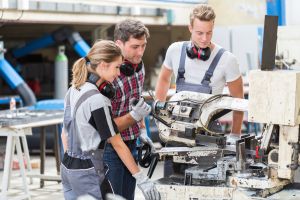 Eine Sch&uuml;lerin und ein Sch&uuml;ler stehen mit einem Ausbilder an einer gro&szlig;en Metallmaschine. Der Ausbilder zeigt ihnen einen Arbeitsschritt.